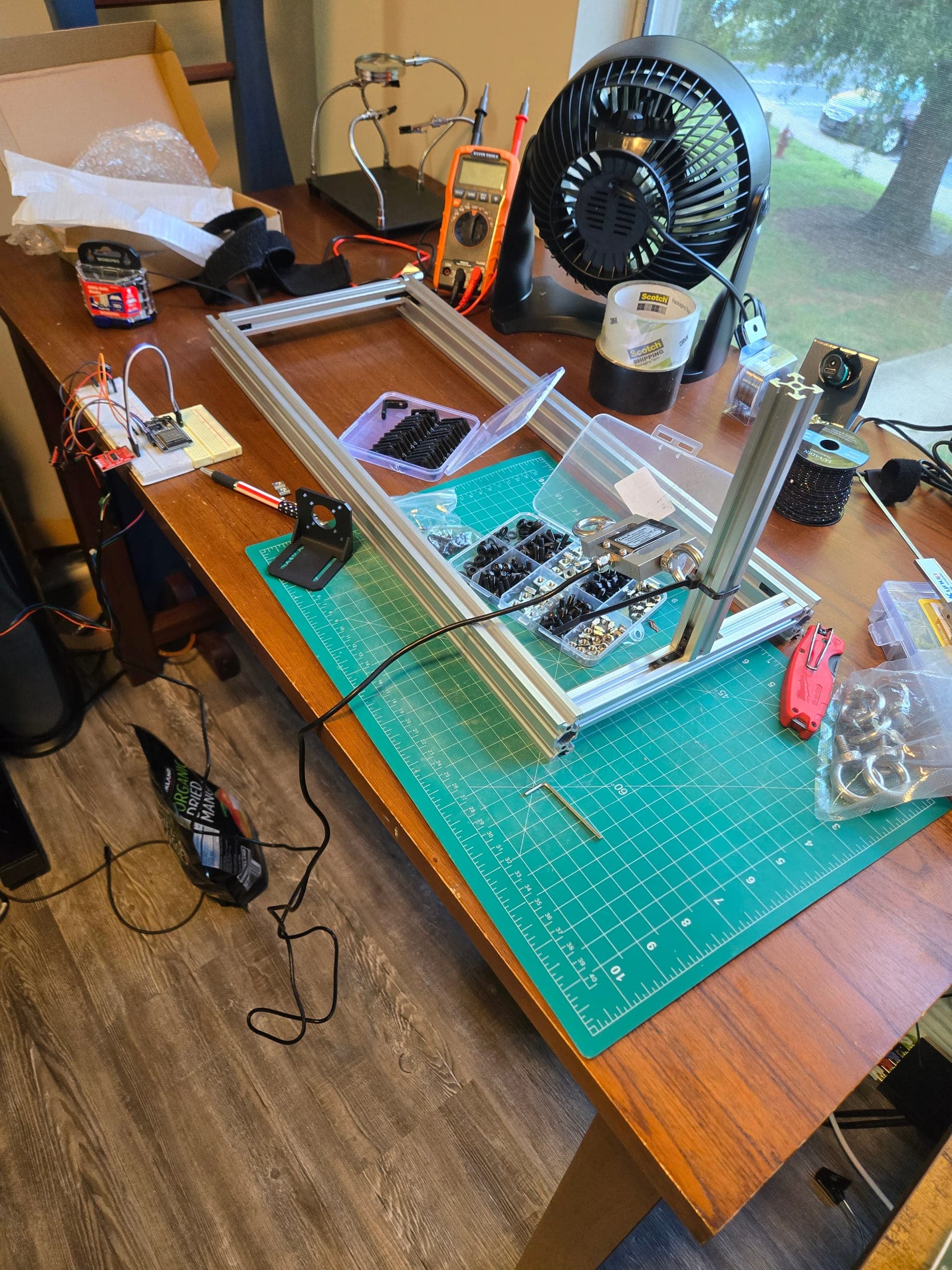 An aluminum extrusion frame under assembly on a green cutting mat, with a vertical post at one corner. An S-shaped load cell hangs from the post on an eye bolt; a black motor bracket lies loose on the mat beside boxes of fasteners.