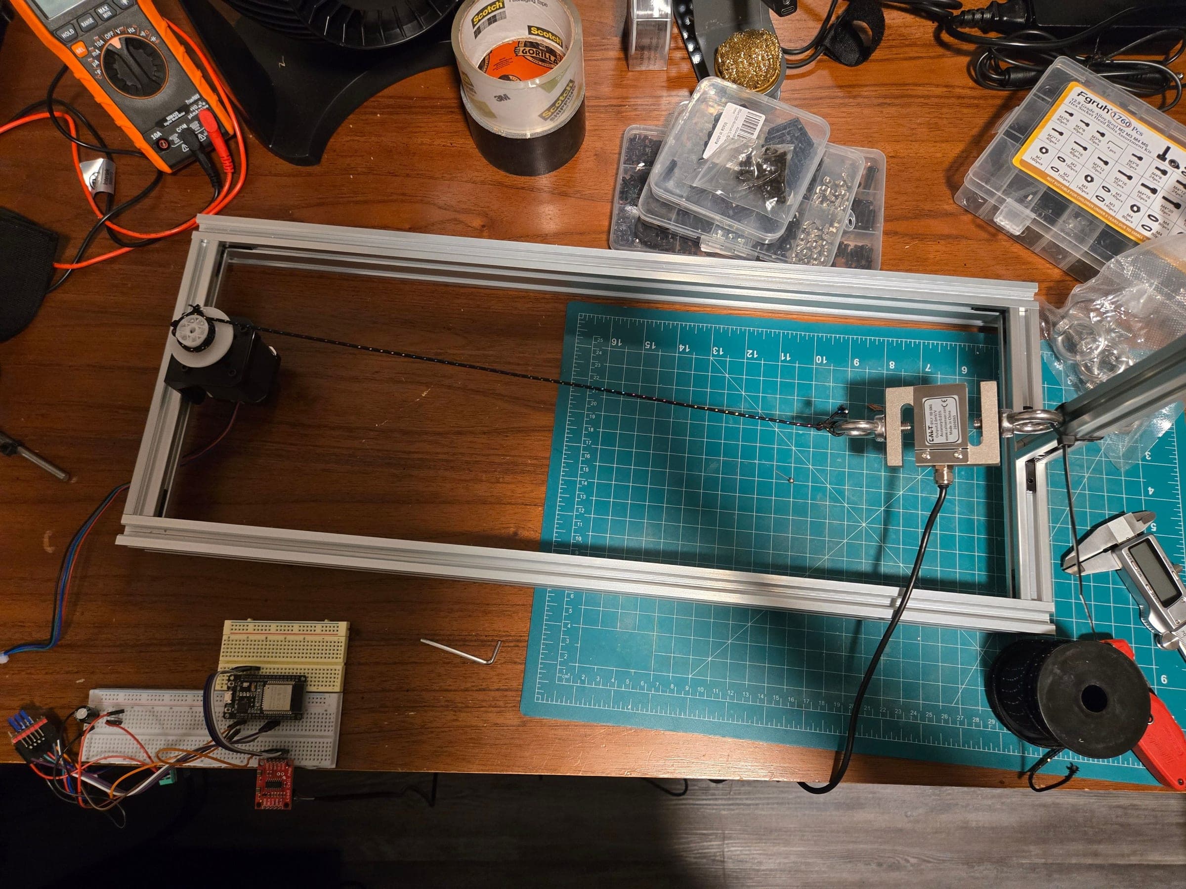 Overhead view of a rectangular aluminum extrusion frame on a desk. A stepper motor with a white printed spool sits at the left end; a braided cord runs across the frame to a silver S-shaped load cell hung on eye bolts at the right. A breadboard with a microcontroller and a red amplifier board sits below the frame.