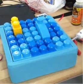 A light-blue square platform covered in a grid of large round blue studs, with a yellow block and a blue block placed on top. The platform sits on a workbench.