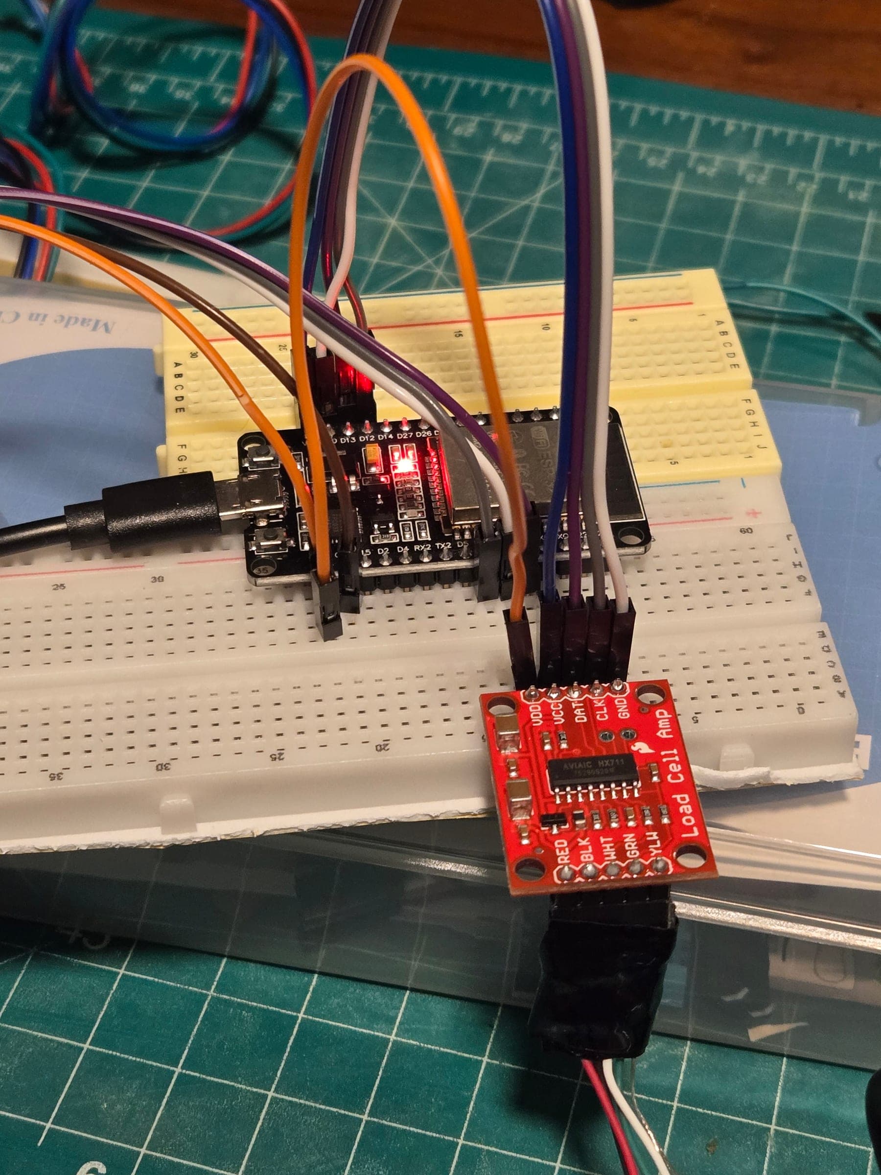 Close-up of a black ESP32 development board on a breadboard with colored jumper wires, connected to a small red SparkFun HX711 load-cell amplifier board.