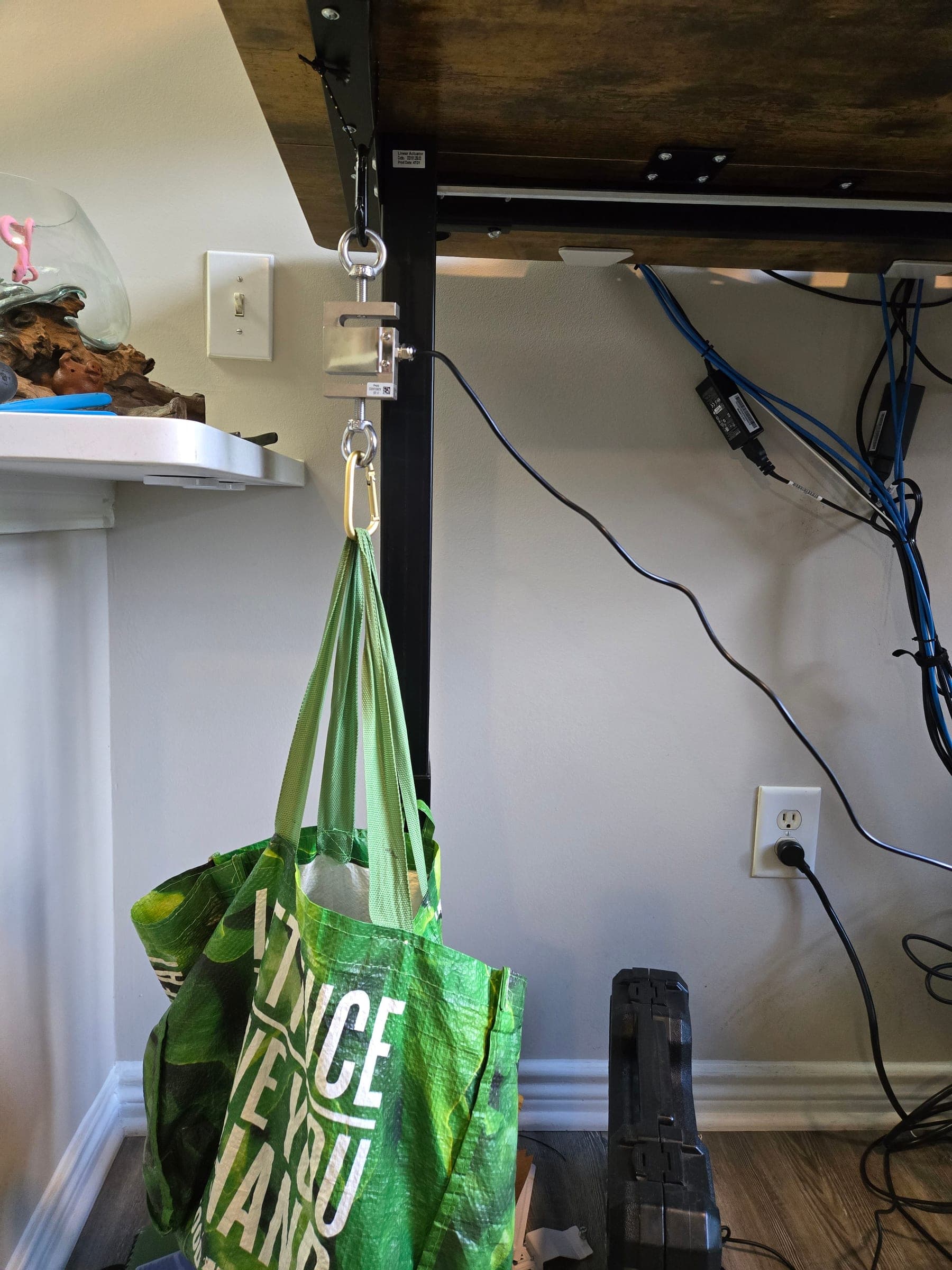 An S-shaped load cell hanging from the underside of a desk by a cord and carabiner, with its signal cable running off to the side and a full reusable shopping bag hanging from a second carabiner below it.