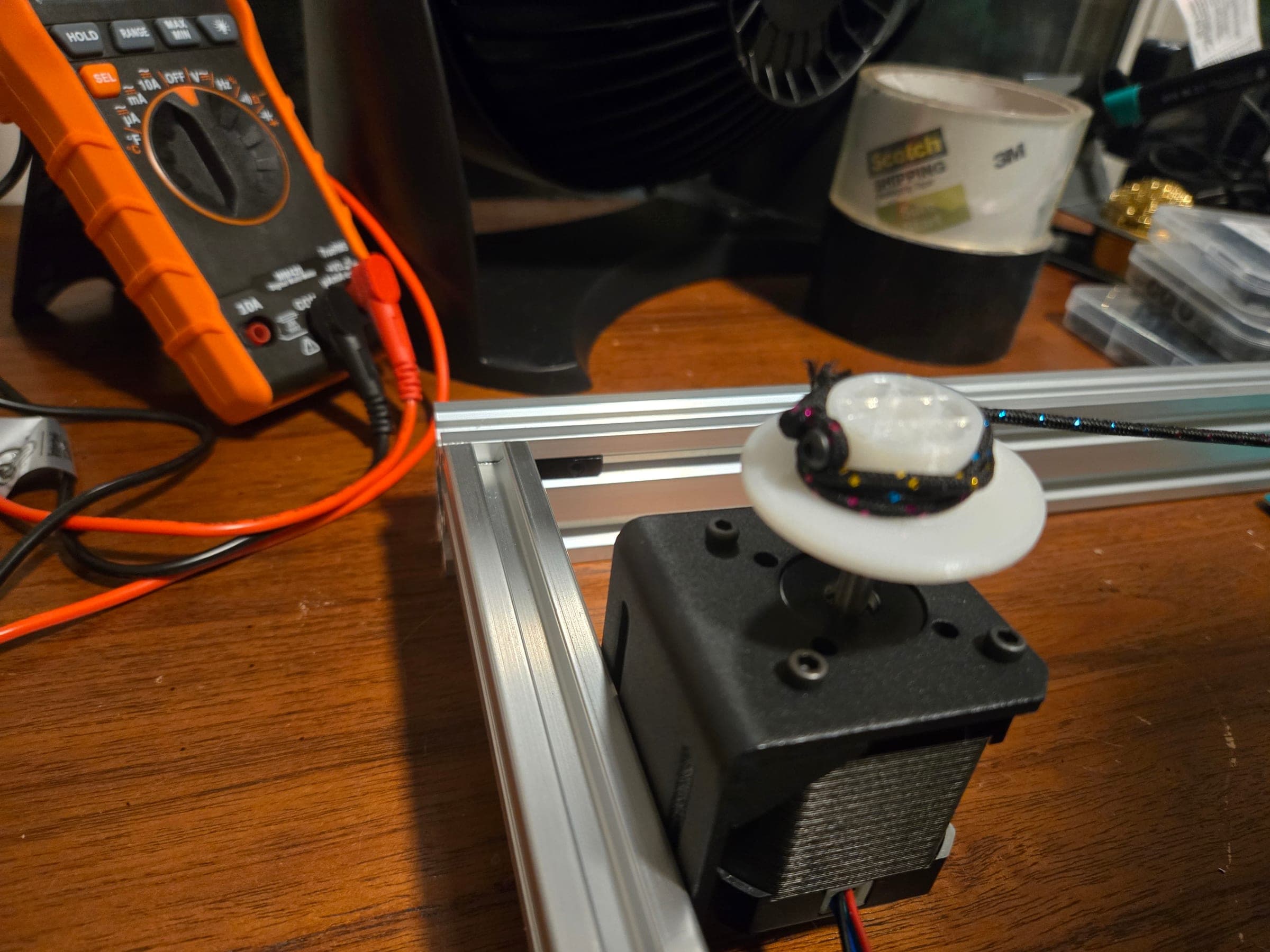 Close-up of a black stepper motor in a black L-shaped bracket standing against an aluminum extrusion, with a white 3D-printed flanged spool on its shaft and braided cord wound on the hub.