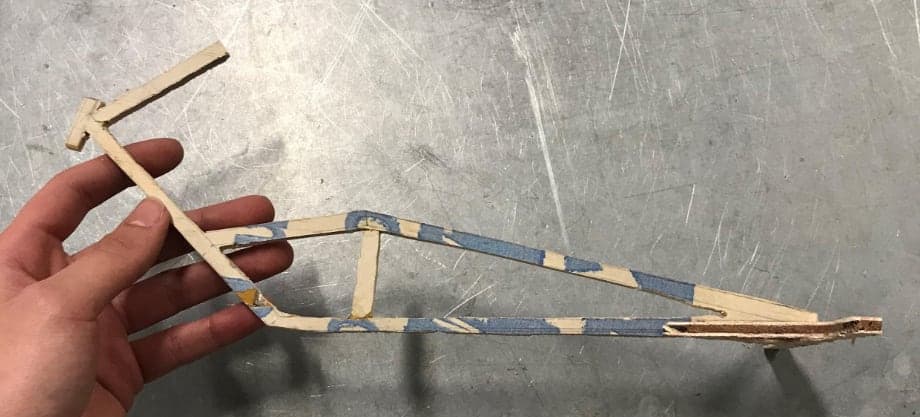 A hand holding a small frame model built from wooden craft sticks, held together with tape.