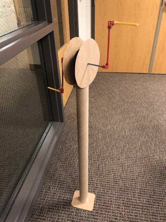 A tabletop model of a hand-crank assembly: a cardboard disk on a post with a red crank arm and yellow handles.