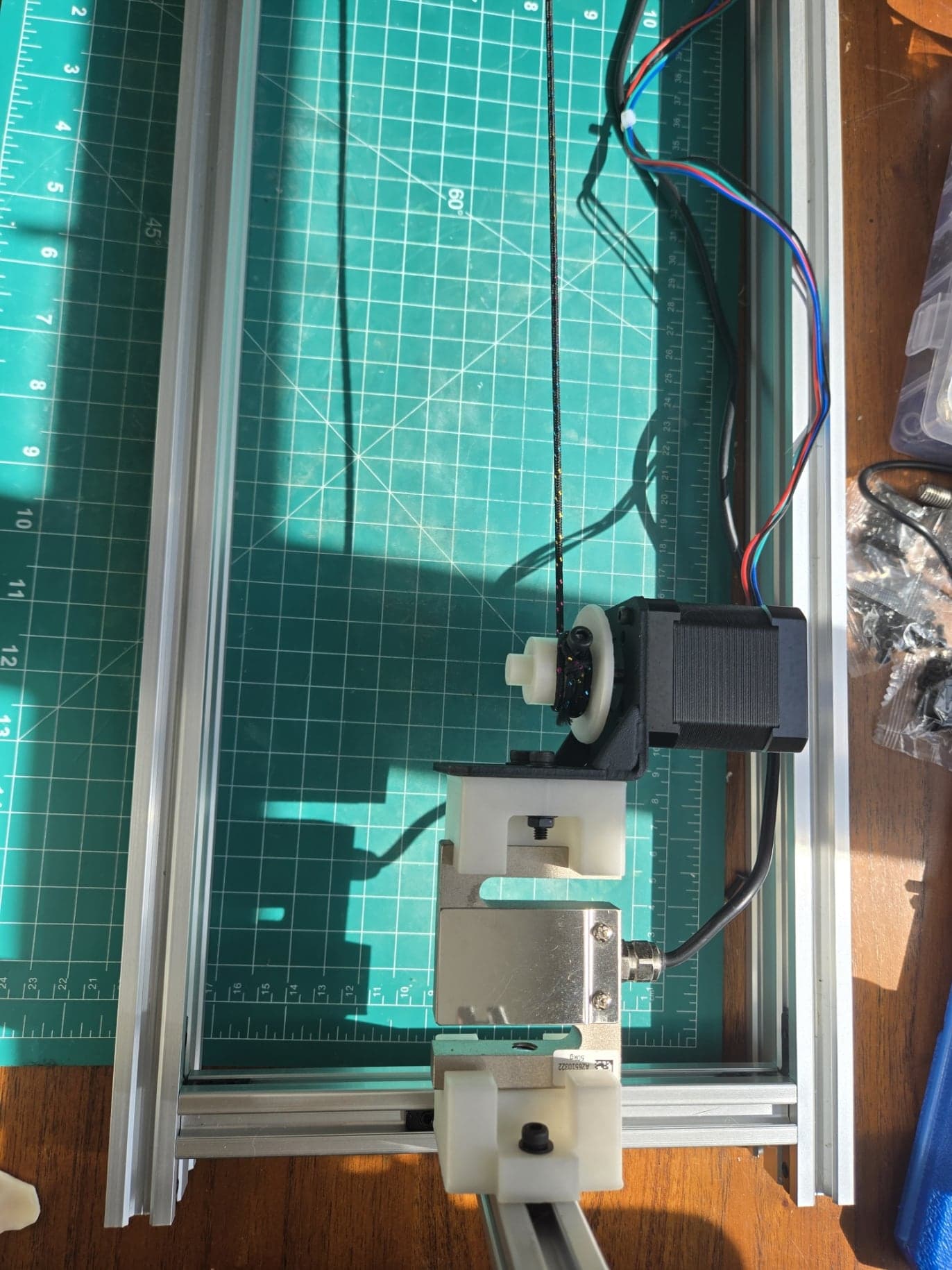 Overhead view of the built fixture on a cutting mat inside an aluminum extrusion frame. A stepper motor with a white printed spool sits in a black L-bracket; the bracket is bolted to a white printed adapter, then a silver S-shaped load cell, then a second white printed adapter fixed to the extrusion. A braided cord runs from the spool up the length of the frame.