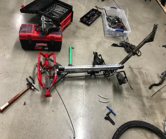 A bare bicycle frame with red rear section and a separate fork lying on a shop floor beside tools and a red toolbox.
