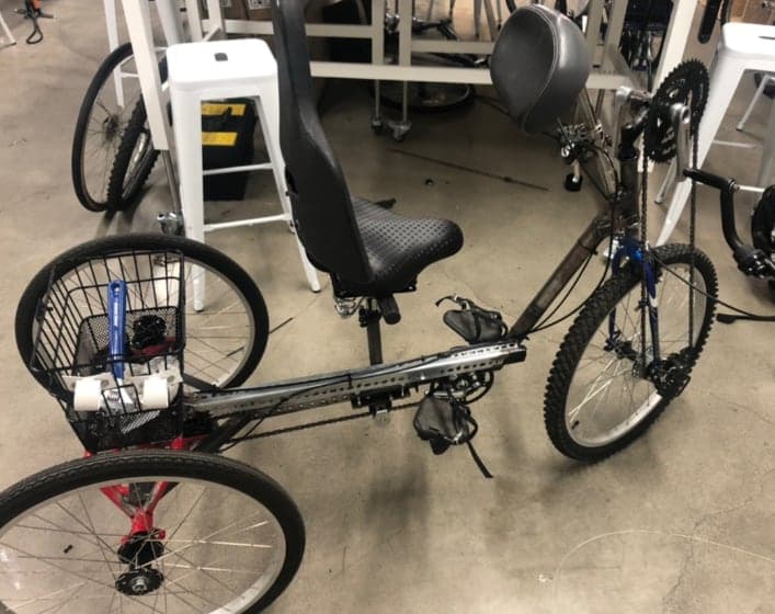 A three-wheeled vehicle in a workshop with a tall padded seat back, a wire basket at the rear, two rear wheels, and a front hand crank.