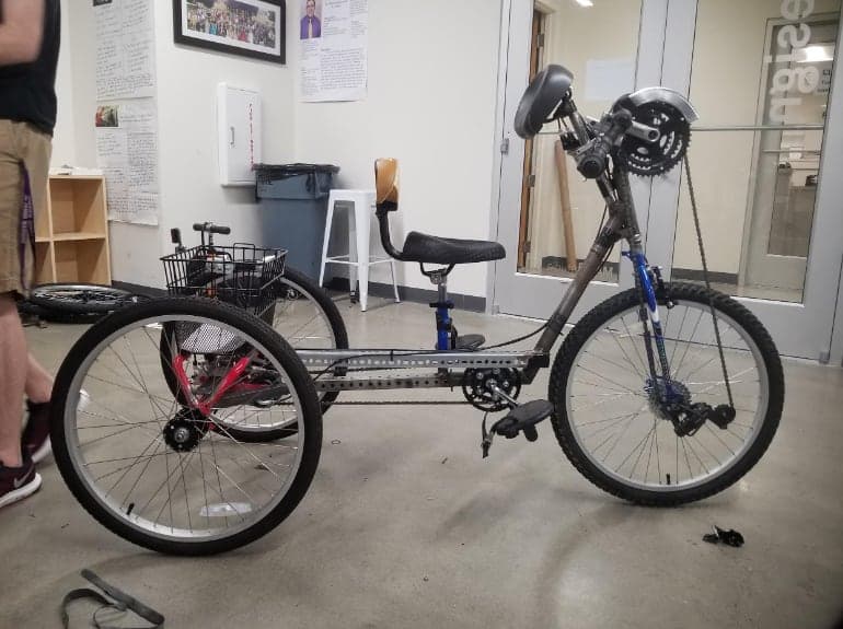 Side view of the finished three-wheeled vehicle in a workshop: steel tube frame, a seat with a short back, a rear basket, foot pedals mid-frame, and a raised hand crank with a chain to the front wheel.