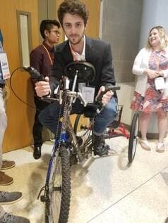 A person in a blazer sits on a three-wheeled hand- and foot-powered bicycle with a tall front hand crank, at an indoor event; other attendees with lanyards stand in the background.