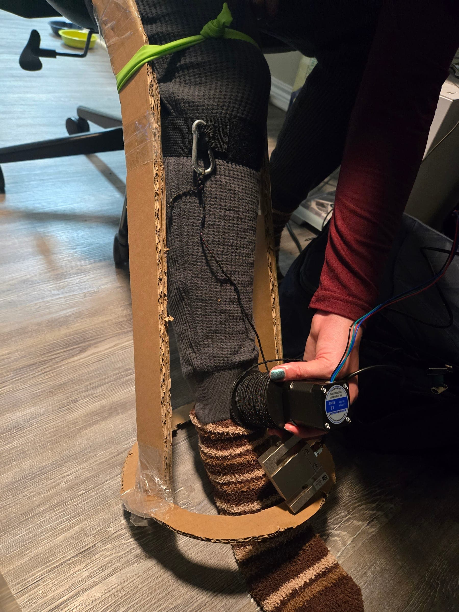 Front view of the cardboard mockup on a leg: a black strap with a metal carabiner below the knee, a braided cord running down the shin, and a hand holding the motor, cord reel, and metal load cell at the cardboard ankle ring.