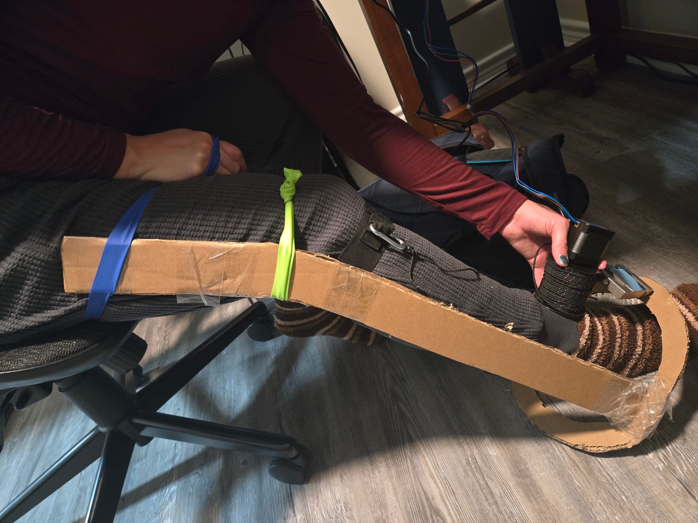 A seated person's leg with a long strip of cardboard taped along the outside from thigh to foot and joined to a cardboard ring at the ankle. A hand holds a stepper motor with a reel of braided cord and a small metal block near the ankle ring.