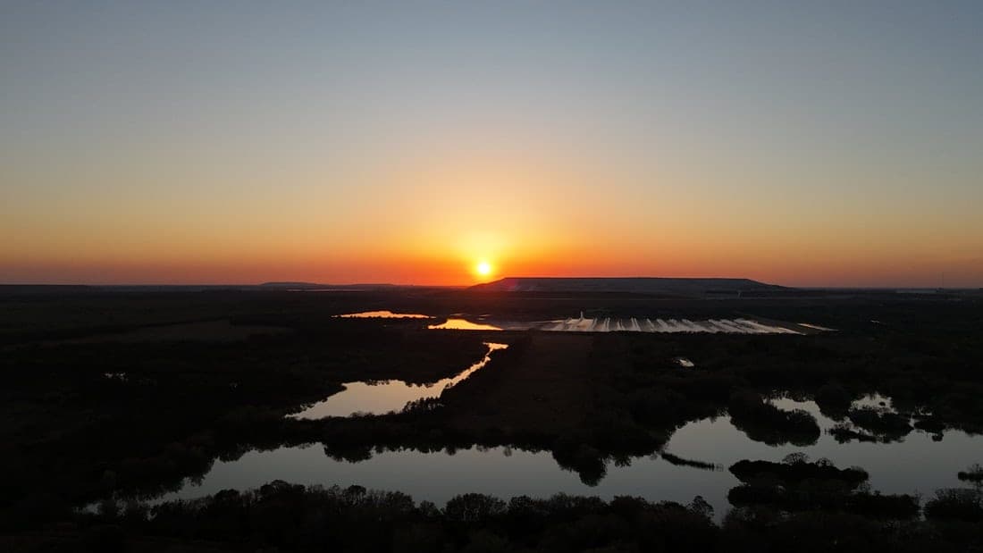 Aerial view of the sun setting on the horizon over flat wetlands with winding channels of water reflecting the sky.