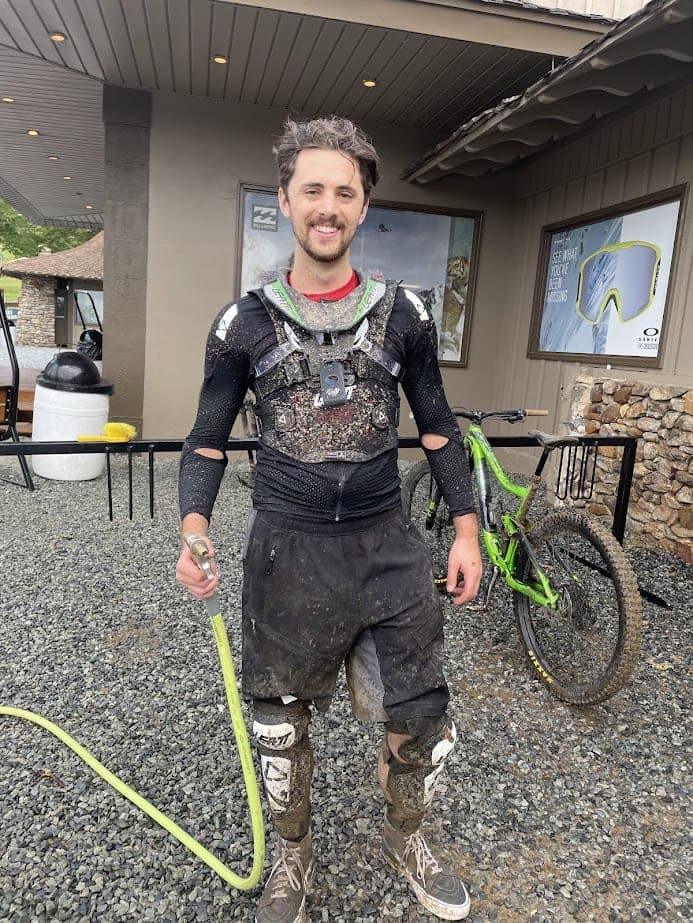 A mountain biker in mud-spattered mountain-bike armor and a chest-mounted camera, standing in front of a bike rack with a green full-suspension mountain bike.