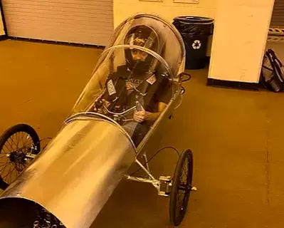 A driver in a helmet sits in a narrow three-wheeled prototype vehicle with a polished aluminum body and a clear canopy, parked indoors.